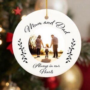 May include: White ceramic Christmas ornament with a family photo inside. The ornament has the words "Mum and Dad" and "Always in our Hearts" in script. The ornament hangs from a gold string, with a Christmas tree in the background.