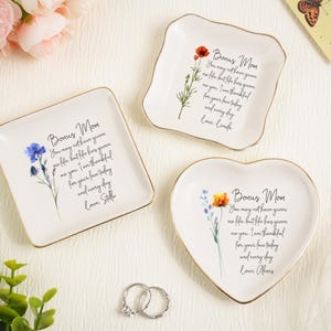 Peut inclure: Trois petits plats à bijoux en céramique blanche avec des bords dorés. L'un est carré, l'autre en forme de cœur et le troisième est un carré ondulé. Chacun a un motif floral et les mots "Bonus Mom" et un message personnalisé.