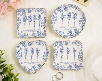 Puede incluir: Cuatro platos decorativos de cerámica con un diseño floral azul y ribete dorado. Cada plato tiene una forma diferente: cuadrado, redondo, corazón y un cuadrado festoneado. El texto "IF BESTIES WERE FLOWERS I'D PICK YOU" está impreso en cada plato.