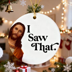May include: A round white Christmas ornament with the text "I Saw That." The ornament features an image of Jesus Christ on the left side. The ornament is hanging from a gold string, with a Christmas tree and lights in the background.