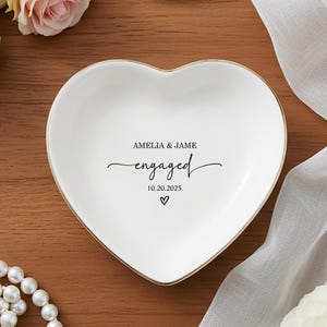 May include: White heart-shaped ceramic ring dish with gold trim. The dish is inscribed with "AMELIA & JAME engaged 10.20.2025" in black script. A pearl necklace and engagement ring are also visible.