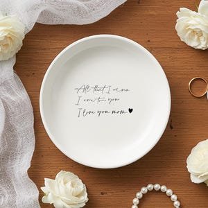 Puede incluir: Un plato de cerámica blanco con la inscripción "All that I am, I owe to you, I love you mom" en escritura negra. El plato está rodeado de rosas blancas, joyas y un paño blanco sobre una superficie de madera.