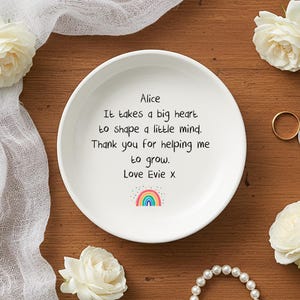 Op de afbeelding: Witte keramische schaal met een handgeschreven boodschap: "Alice, Er is een groot hart nodig om een kleine geest te vormen. Bedankt dat je me hebt geholpen te groeien. Love Evie x." Een regenboogafbeelding staat onder de tekst. Witte rozen en sieraden.