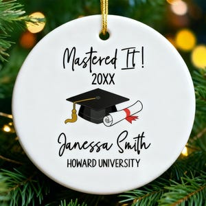 以下が含まれることがあります： 金色の吊り紐が付いた白いセラミック卒業オーナメント。「Mastered It!」の文字と「20XX」の年、卒業キャップと卒業証書が特徴です。下には「Janessa Smith」と「HOWARD UNIVERSITY」の名前があります。