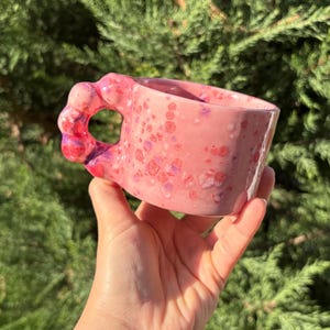 May include: A pink ceramic mug with a distinctive handle, speckled with darker pink and purple spots. The mug is held in a hand, set against a backdrop of green foliage. The mug is likely handmade.