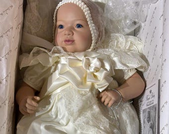 May include: A porcelain doll in a box, wearing a cream satin gown with lace trim and a matching bonnet. The doll has blue eyes and a realistic appearance. The box is lined with tissue paper.