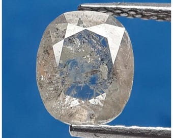 May include: A clear, oval-shaped gemstone with a faceted top, held by silver tweezers against a blue background. The gem has internal inclusions and a slightly cloudy appearance, showcasing its natural characteristics.