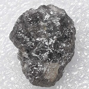 Op de afbeelding: Een ruwe, ongeslepen, donkergrijze diamant met een getextureerd oppervlak. De natuurlijke edelsteen heeft een kristallijn uiterlijk en reflecteert licht, wat de ruwe, ongepolijste staat laat zien. De diamant staat tegen een witte achtergrond.