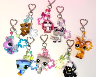 May include: A collection of seven keychains featuring various plastic animal figures with large eyes. Each keychain has a silver heart-shaped clasp, colorful beads, small bows, and star-shaped charms in shades of blue, pink, yellow, and green.