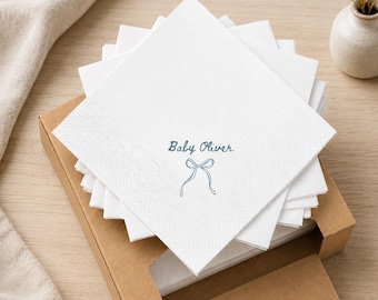 May include: A stack of square white paper napkins featuring the blue printed text Baby Oliver above a simple blue line drawing of a bow. The napkins are housed in an open brown cardboard box, displaying a textured border on the paper edges.