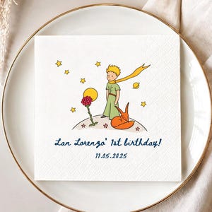 May include: A white square paper napkin featuring a colorful illustration of a young boy with blonde hair, a red rose, and an orange fox on a small planet with stars. The text reads Lan Lorenzo' 1st birthday! and the date 11.05.2025 in blue.