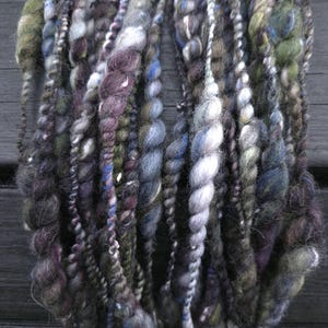 May include: Close-up of a bundle of hand-spun yarn in shades of gray, purple, blue, and green. The yarn has a thick, textured appearance with varying thicknesses and twists, suitable for fiber art projects.