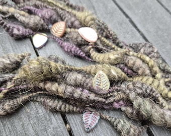 May include: Close-up of handmade dreadlocks in shades of brown, purple, and olive green. Several leaf-shaped beads in various colors are interspersed among the dreadlocks. The dreadlocks are arranged on a wooden surface.