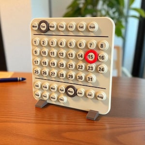 Peut inclure: Un calendrier perpétuel beige avec une grille de jours et de mois numérotés. Le jour 15 est mis en évidence en rouge. Le calendrier a une bordure marron pour le mois et le jour de la semaine. Le calendrier est sur une table en bois.