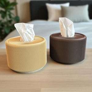 Op de afbeelding: Twee cilindrische tissueboxhouders. Eén is lichtgeel, de andere bruin. Beide hebben een geribbeld ontwerp en een witte tissue die uit de bovenkant steekt. De tissueboxen staan op een houten oppervlak.