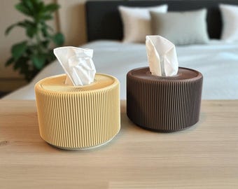 Könnte beinhalten: Zwei zylindrische Taschentuchboxen. Eine ist hellgelb, die andere braun. Beide haben ein geripptes Design und ein weißes Taschentuch, das oben herausragt. Die Taschentuchboxen stehen auf einer Holzoberfläche.