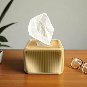 Pode incluir: Uma caixa de lenços de papel amarelo claro com detalhes canelados verticais. Os lenços brancos são puxados de cima. A caixa está sobre uma superfície de madeira, com um par de óculos à direita e uma planta em vaso à esquerda.