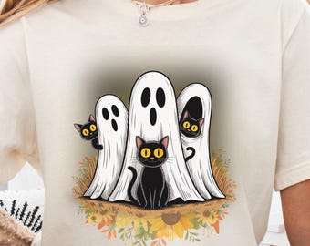 May include: Cream-colored t-shirt featuring a Halloween design. The graphic includes three white ghosts with black eyes and a black cat. The design is set on a bed of flowers and grass. Text at the bottom reads "PNG File - Digital Download Only (No Physical Item)".