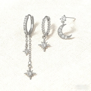 Puede incluir: Un conjunto de tres pendientes plateados. Uno es un aro con una cadena y tres colgantes de estrella. Otro es un aro con un colgante de estrella. El tercero es un pendiente de botón con un colgante de luna creciente y una estrella.