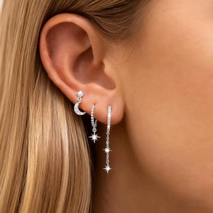 May include: Close-up of an ear with three silver earrings. One earring has a crescent moon and star, the others have dangling stars and small, sparkling stones. The earrings are set against a blurred background. The earrings are made of silver.