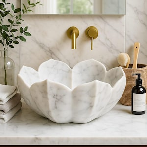 May include: A white marble lotus-shaped sink with gold taps. The sink is the focal point, set against a marble wall and countertop. A mirror, greenery, and a woven basket with toiletries complete the bathroom scene.