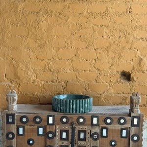 May include: A decorative wooden cabinet with intricate metal and mirror accents, topped with a ribbed, teal-colored bowl. The cabinet is set against a textured, ochre-colored brick wall, creating a rustic and artistic display.