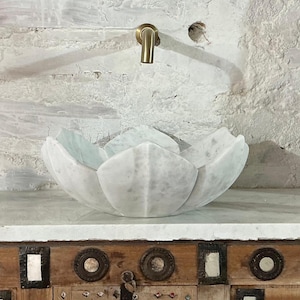 May include: A white marble lotus flower sink sits on a wooden cabinet with decorative metal accents. A brass tap is mounted on the textured white wall behind the sink. The cabinet has carved wooden details on each side.