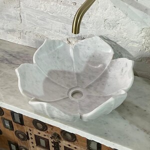 May include: A white marble flower-shaped sink with a gold tap against a textured white wall. The sink is set on a marble countertop above a wooden cabinet with decorative metal accents. The tap is curved.