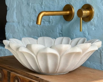 May include: A white marble lotus flower-shaped sink sits atop a wooden cabinet. The sink is paired with gold-colored faucet fixtures against a light blue painted wall. A wooden framed mirror is above the faucet.