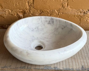 May include: Oval-shaped white marble sink with a central drain hole. The sink is set on a wooden cabinet with decorative metal accents. The background features a textured, orange-brown brick wall.