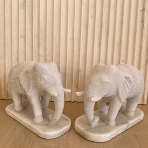 May include: Two white marble elephant sculptures. Each elephant has detailed carvings and stands on a rectangular base. The elephants have trunks, tusks, and large ears. The background is a light-colored, vertically-lined wall.