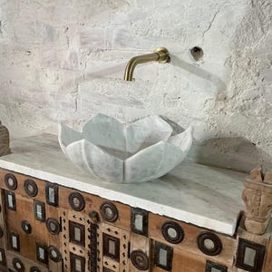 May include: A marble lotus-shaped sink sits on a wooden cabinet with metal detailing. A brass tap is mounted on the white brick wall. The sink and cabinet create a unique bathroom design.