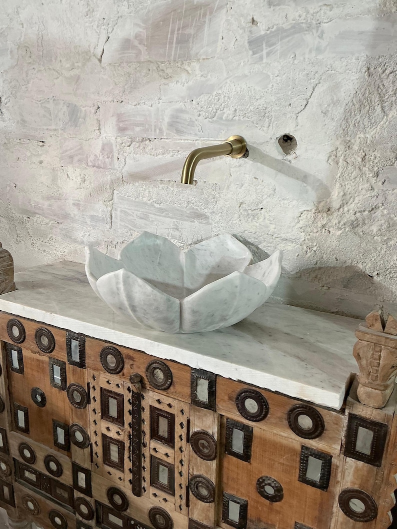 May include: A marble lotus-shaped sink sits on a wooden cabinet with metal detailing. A brass tap is mounted on the white brick wall. The sink and cabinet create a unique bathroom design.