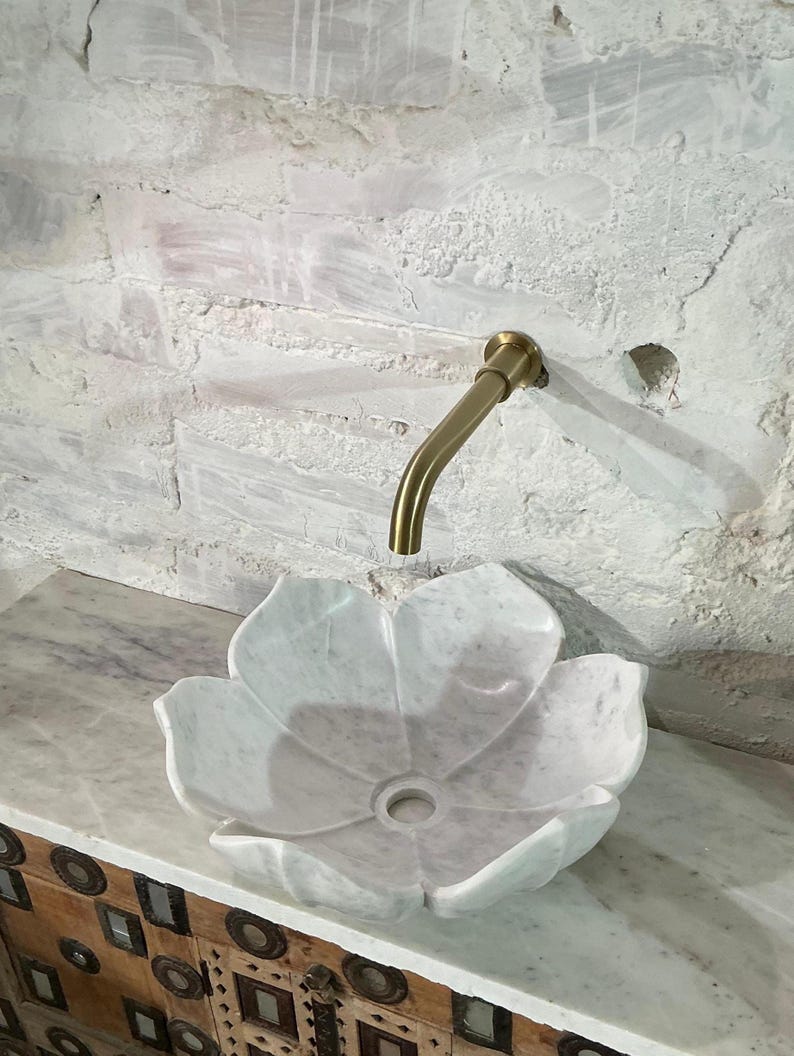 May include: A white marble flower-shaped sink with a gold tap against a textured white wall. The sink is set on a marble countertop above a wooden cabinet with decorative metal accents. The tap is curved.