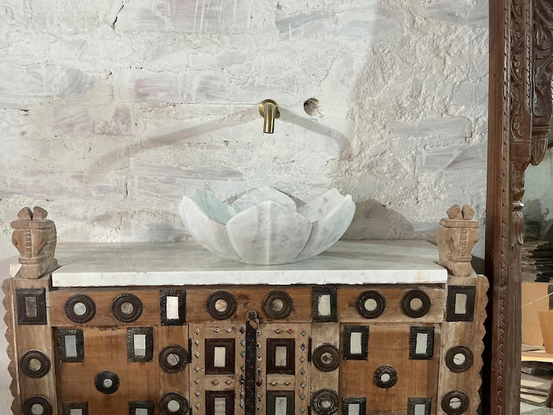 May include: A white marble lotus flower sink sits on a wooden cabinet with decorative metal accents. A brass tap is mounted on the textured white wall behind the sink. The cabinet has carved wooden details on each side.