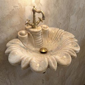May include: A decorative, shell-shaped bathroom sink made of light-colored stone. The sink has a brass faucet and drain. The sink is mounted on a textured wall. The sink's design is reminiscent of a seashell.