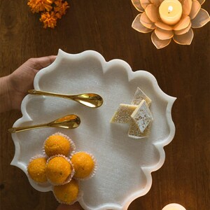 May include: A white marble serving tray with a scalloped edge, holding golden spoons, orange sweets, and diamond-shaped treats. The tray is held by hands, with marigold flowers and a lotus-shaped candle holder in the background.
