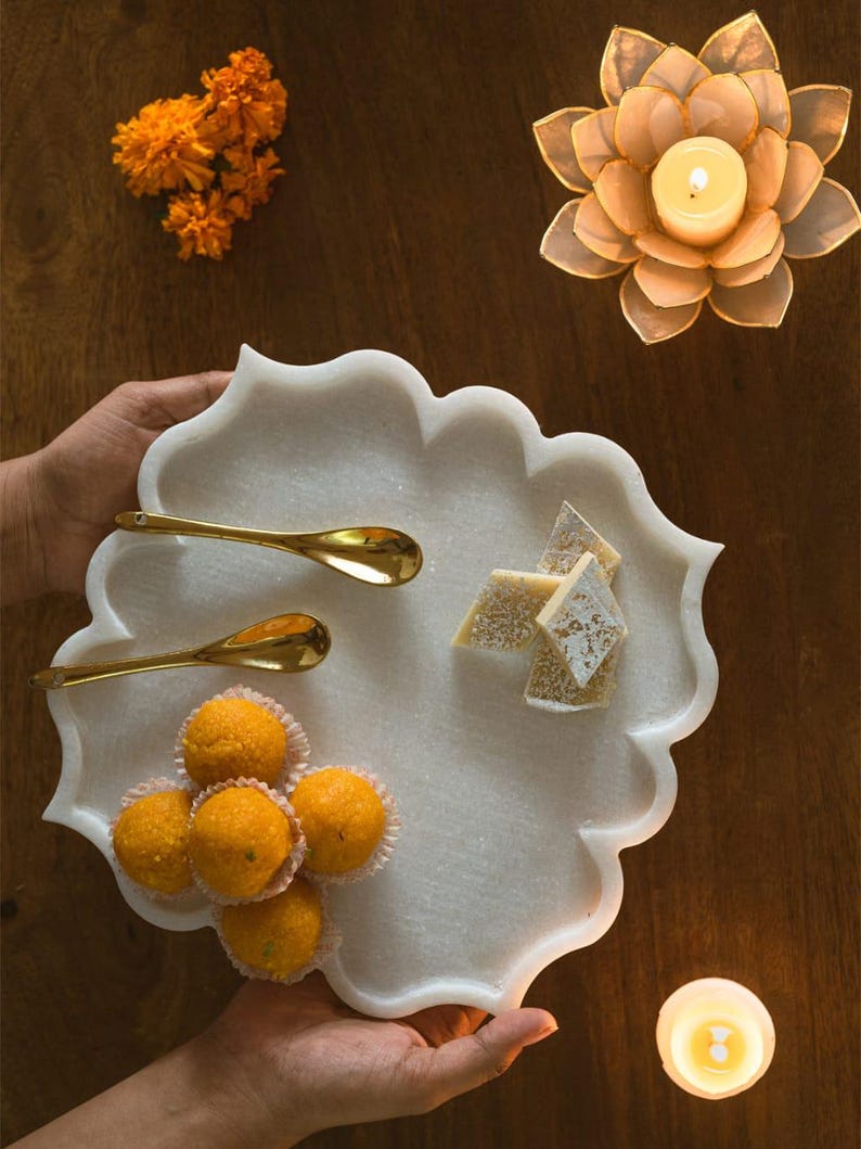 May include: A white marble serving tray with a scalloped edge, holding golden spoons, orange sweets, and diamond-shaped treats. The tray is held by hands, with marigold flowers and a lotus-shaped candle holder in the background.