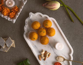 May include: A marble serving tray with orange sweets, nuts, and spices is the focal point. A small candle and marigold flowers are in a decorative dish. Lotus buds are on the right. The scene is set on a grey surface.