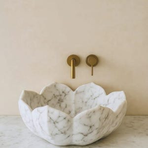 May include: A white marble sink with a flower-petal design. The sink is set against a light beige wall, with gold-coloured faucet fixtures. The sink is made of white marble with grey veining.
