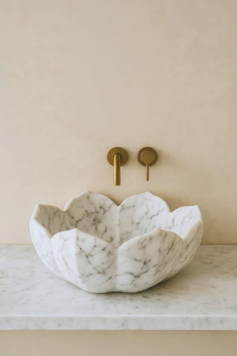 May include: A white marble sink with a flower-petal design. The sink is set against a light beige wall, with gold-coloured faucet fixtures. The sink is made of white marble with grey veining.
