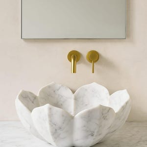 May include: A white marble sink with a flower petal design. The sink is set against a neutral wall with a rectangular mirror and two gold-coloured faucet handles. The sink is a unique bathroom fixture.