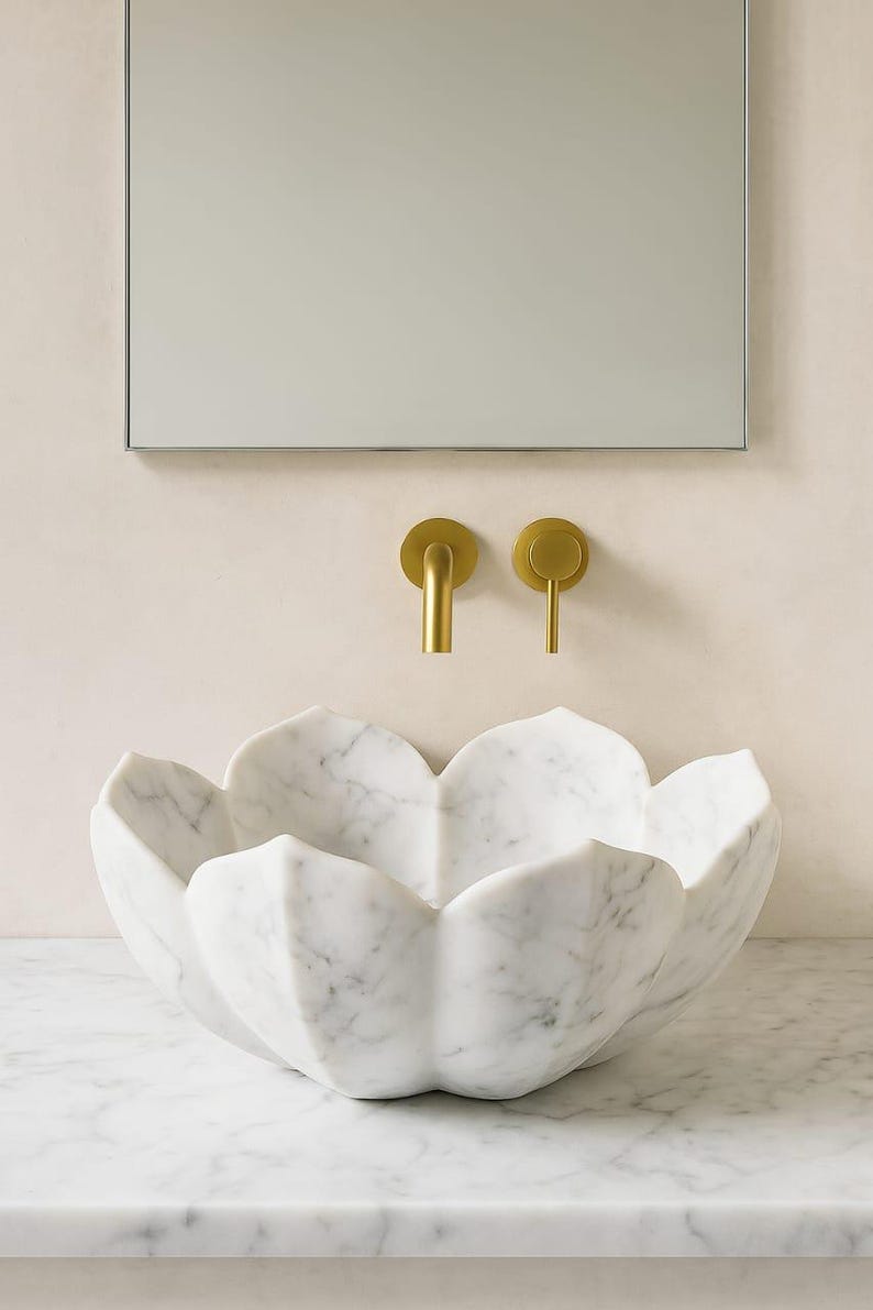 May include: A white marble sink with a flower petal design. The sink is set against a neutral wall with a rectangular mirror and two gold-coloured faucet handles. The sink is a unique bathroom fixture.