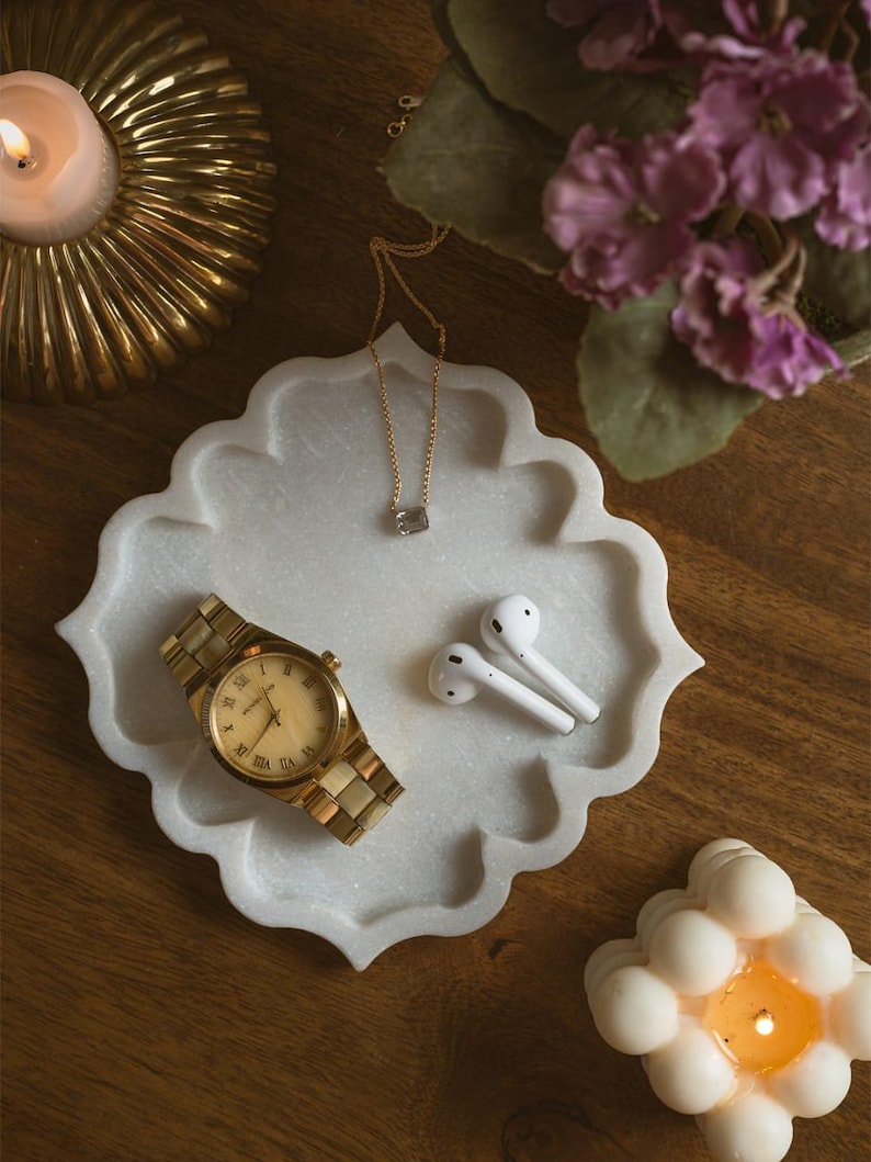 May include: A gold wristwatch with a light-colored face and a gold link band rests on a white marble tray. The tray also holds white earbuds and a gold necklace with a small pendant. A lit candle and purple flowers are in the background.
