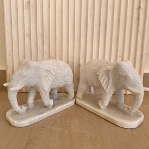 May include: Two white stone elephant sculptures. Each elephant has detailed carvings and is standing on a rectangular base. The elephants have large ears, trunks, and tusks. The background features a textured, light-colored wall.