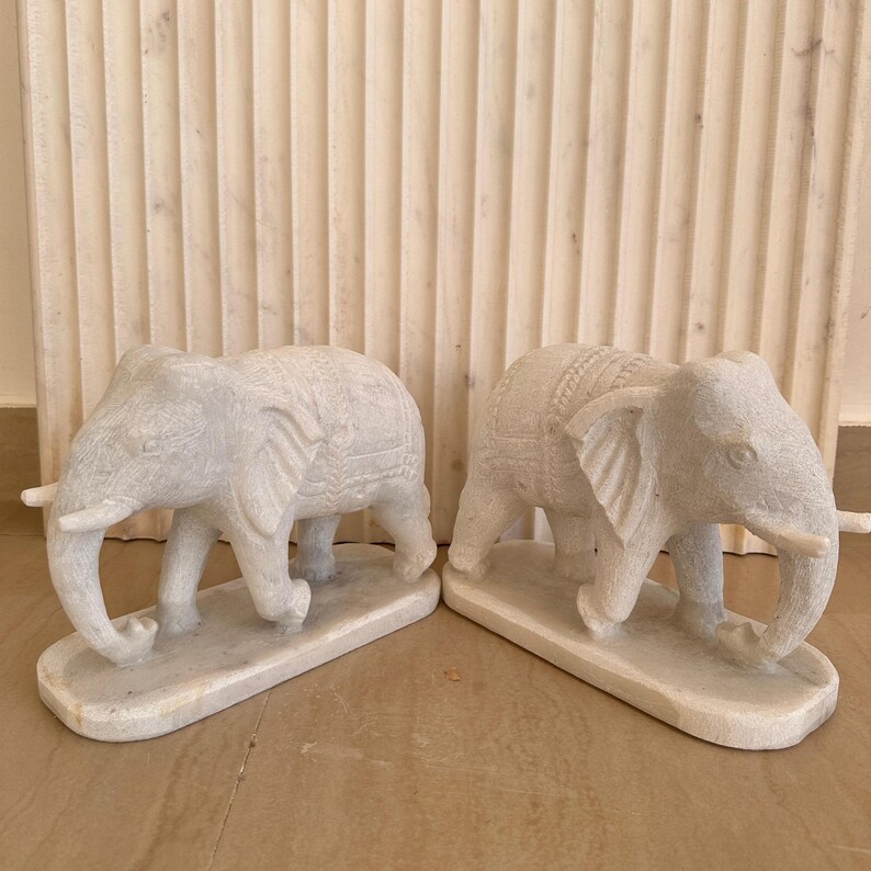 May include: Two white stone elephant sculptures. Each elephant has detailed carvings and is standing on a rectangular base. The elephants have large ears, trunks, and tusks. The background features a textured, light-colored wall.