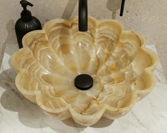 May include: A flower-shaped vessel sink carved from translucent beige and cream-colored stone with natural veining. The basin features a matte black drain plug and is paired with a matching matte black faucet fixture above the center.