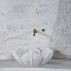 May include: A white marble lotus flower-shaped sink with a gold tap against a white brick wall. The sink is a unique, artistic design, ideal for a modern bathroom. The tap is mounted on the wall above the sink.