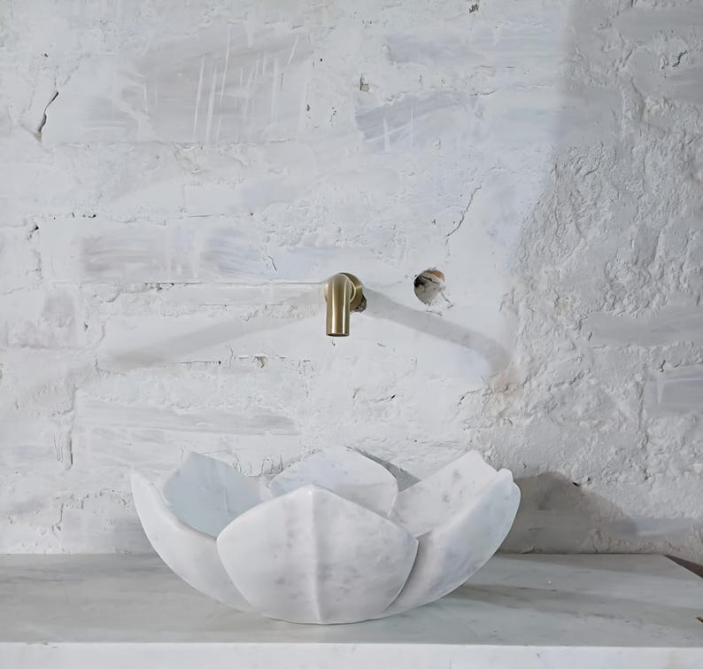 May include: A white marble lotus flower-shaped sink with a gold tap against a white brick wall. The sink is a unique, artistic design, ideal for a modern bathroom. The tap is mounted on the wall above the sink.
