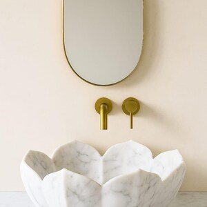 May include: A white marble lotus-shaped sink with a gold tap and matching fittings. An oval gold-framed mirror hangs above the sink. The sink is set against a light beige wall.